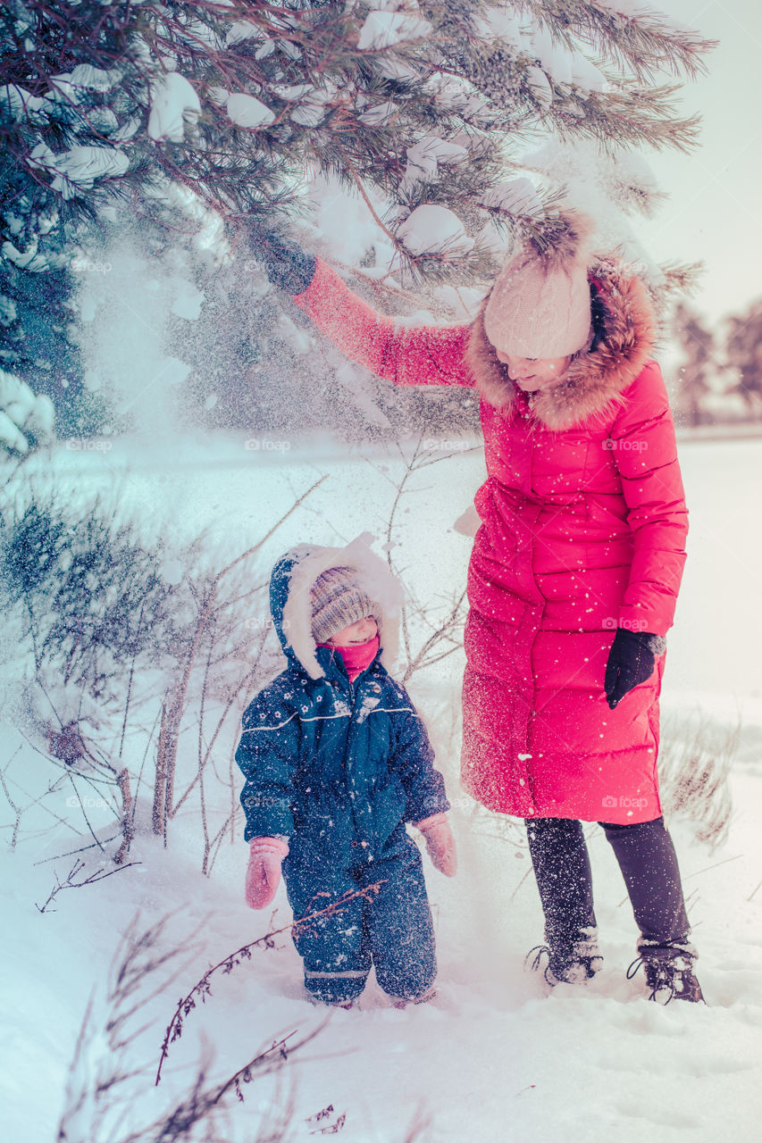 Mother and her little daughter are spending time together outdoors playing with snow. Mother is wearing red winter coat and wool cap, toddler is wearing dark blue snowsuit