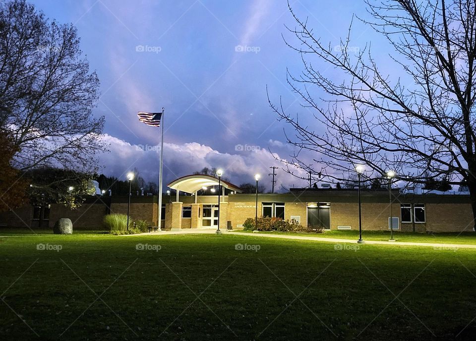 Low puffy clouds at dusk turn pink, purple, and white in a blue sky behind a brick school building with an American flag flapping in the wind on a bright green lawn