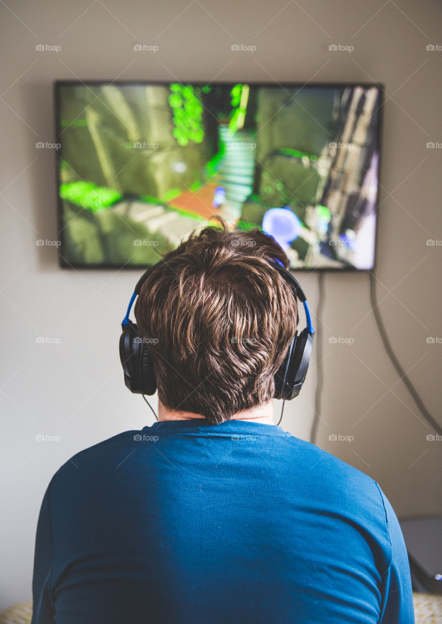 A young man playing video games alone in his room and watching the tv with headphones on
