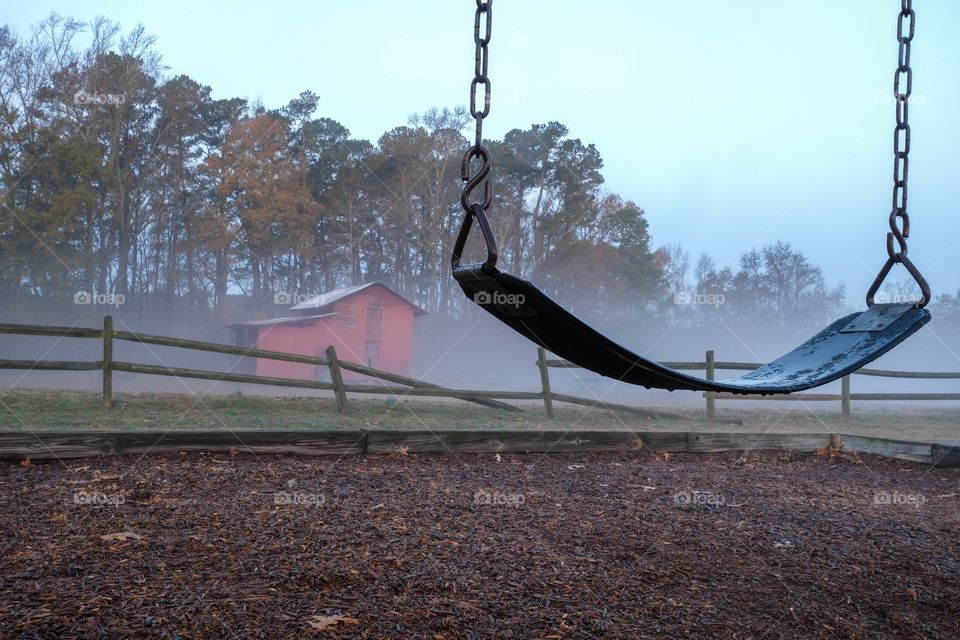 A lonesome melancholic scene of an empty swing with an old red barn and a broken wooden fence on a cool foggy morning. Lake Benson Park in Garner North Carolina. 