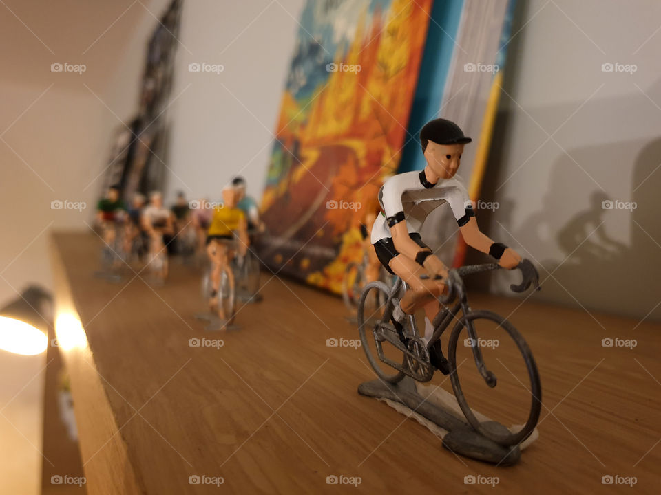 A portrait of some miniature cyclists racing over a wooden bookshelf indoor in a house.