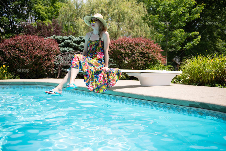 Young millennial woman wearing a dress and flip flops while sitting on the diving board of a swimming pool in the summer