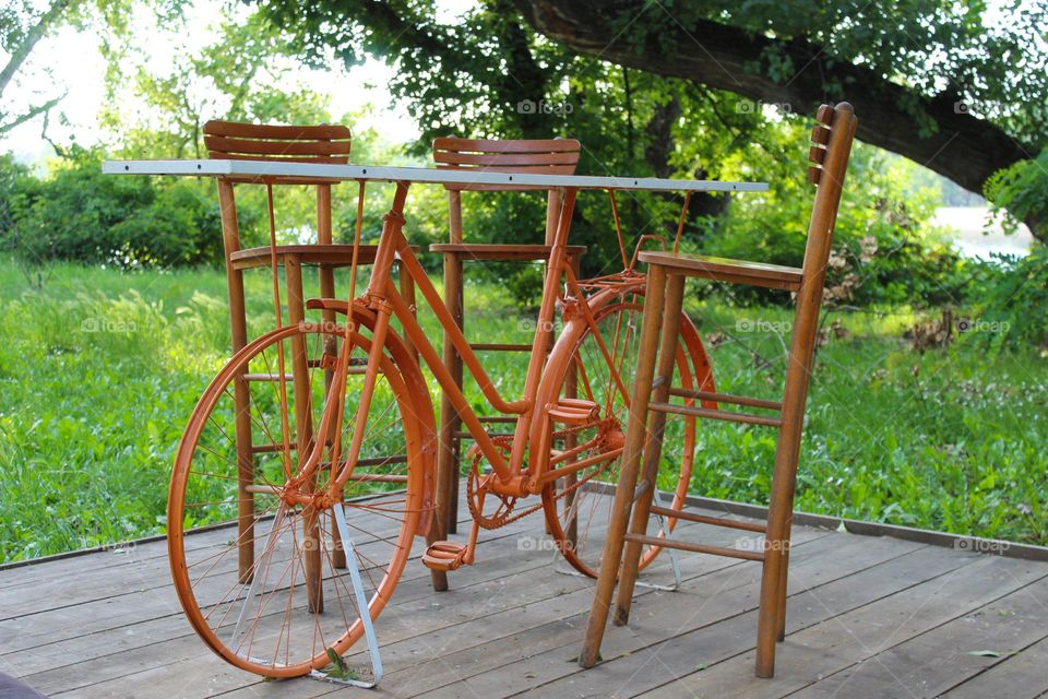 One lonely orange bicycle with a wooden board on the top and two chairs.  Bicycle as a table