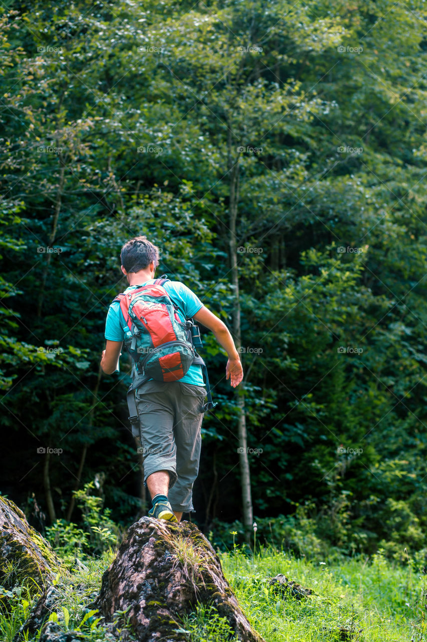 Young wanderer with backpack walking over a rock towards forest. Spends a vacation on wandering with backpack, he is wearing sport summer clothes