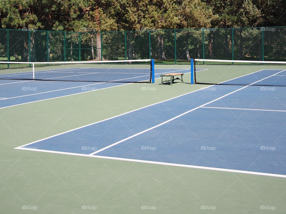 An outdoor blue and green tennis court surrounded by fencing and trees. 