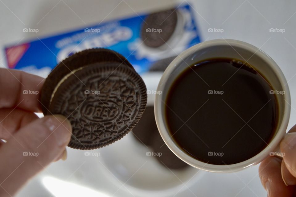 A person holding an Oreo cookie and a cup of coffee