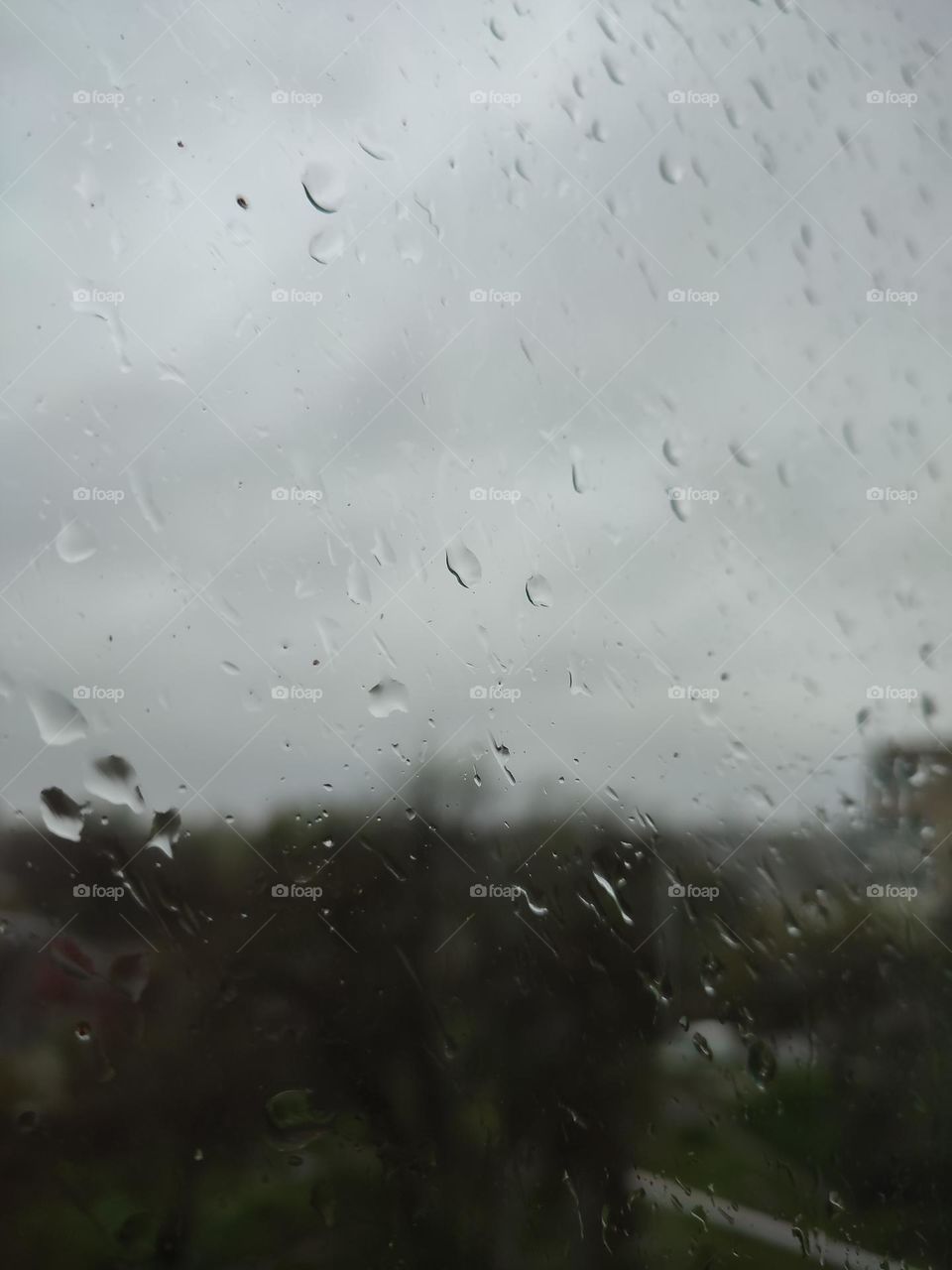 Raining day in the town, view from the window. Raindrops running on the window glass. Blurred skyline, trees, multi storey building at the background. Spring thunderstorm, gray heavy sky. View from the window, wet weather. Rain in the city