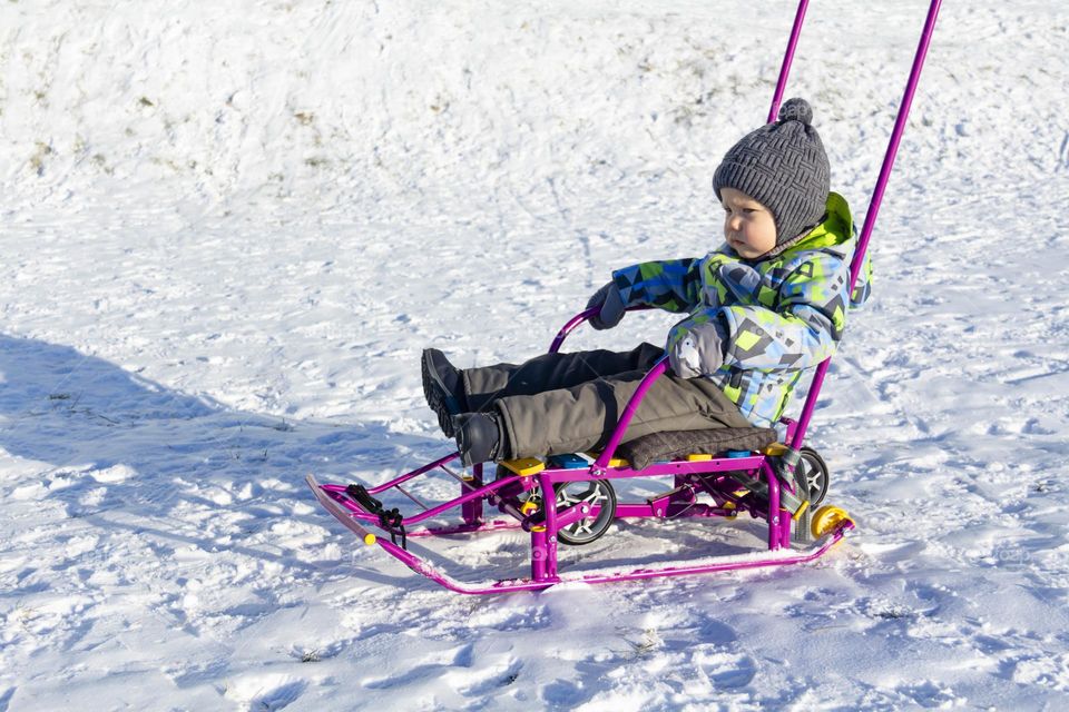 A child with a serious expression on his face in winter clothes jackets, pants, hat and boots in winter on white snow on the street and in the park in nature sledding and playing winter fun.