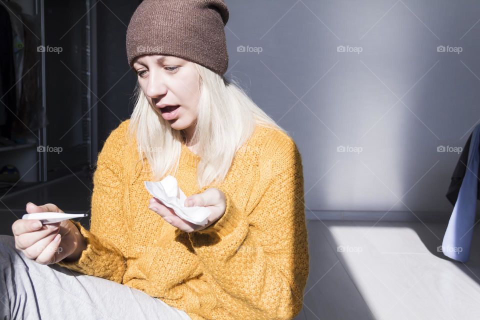 A young girl in a yellow sweater and a brown hat is very surprised at the high temperature on the thermometer with a napkin in her hands.
