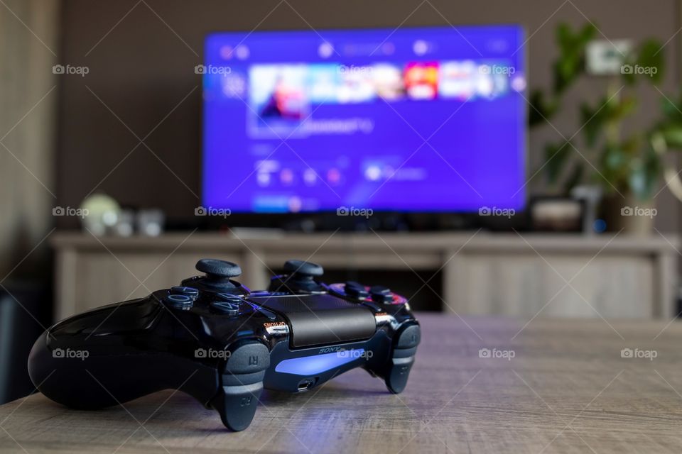 A portrait of a limited edition PlayStation 4 controller, in the blurry background there is a television with the PlayStation homescreen being displayed.