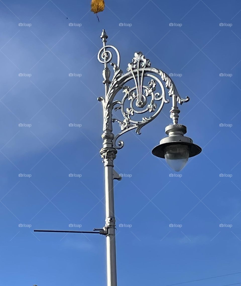 The photo captures an antique-looking street lamp in Dublin, its ornate metalwork reflecting a sense of old-world charm. A single brown leaf hovers in mid-air, caught in a gentle breeze, seemingly making its way over the lamp as if drawn to it.