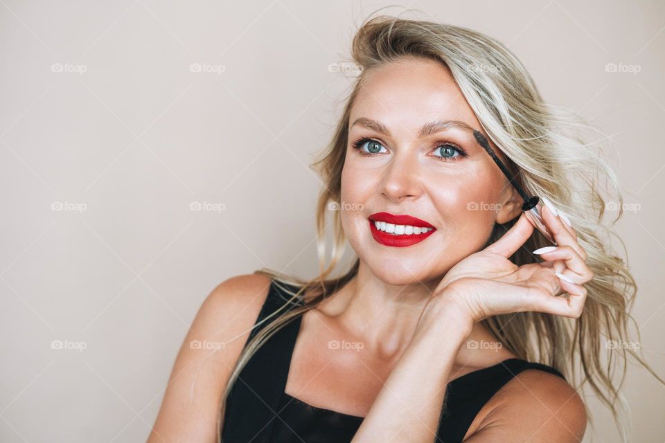 Portrait of gorgeous happy blonde young woman with bright makeup with eyebrow mascara brush in evening dress on beige background