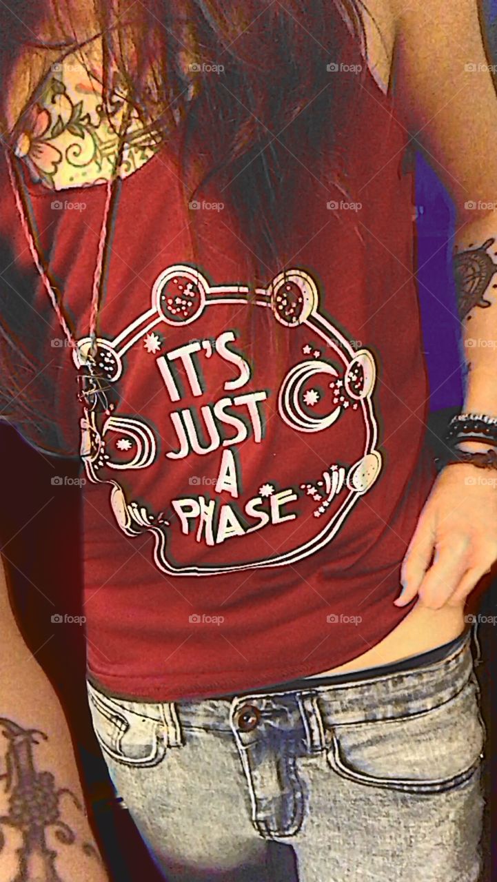 A slender Hispanic model’s torso is visible in a maroon shirt with a moon cycle design that reads “It’s Just A Phase”. beaded bracelets and a hemp rope with an Obsidian stone pendant, visible forearm tattoo, slightly lifting corner of shirt at hip. 