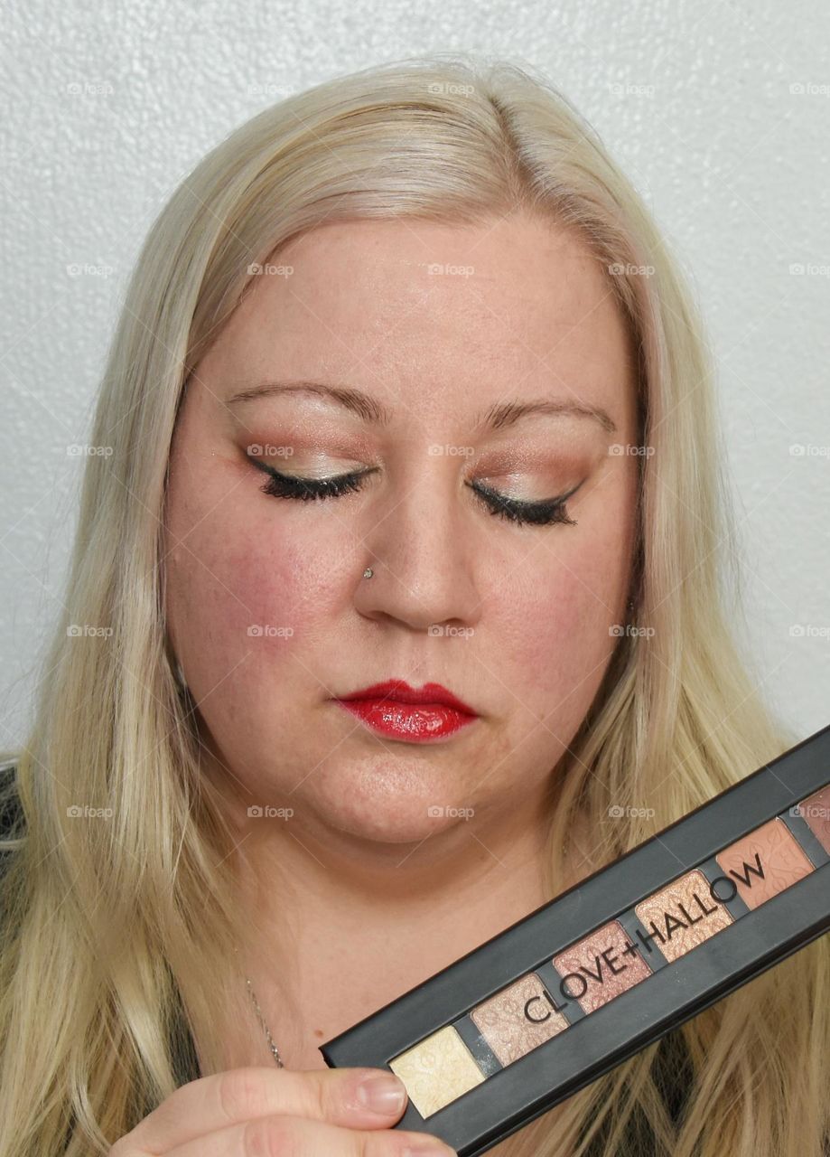 Blonde woman holding eye shadow assortment with eyeshadow on her eye lids 