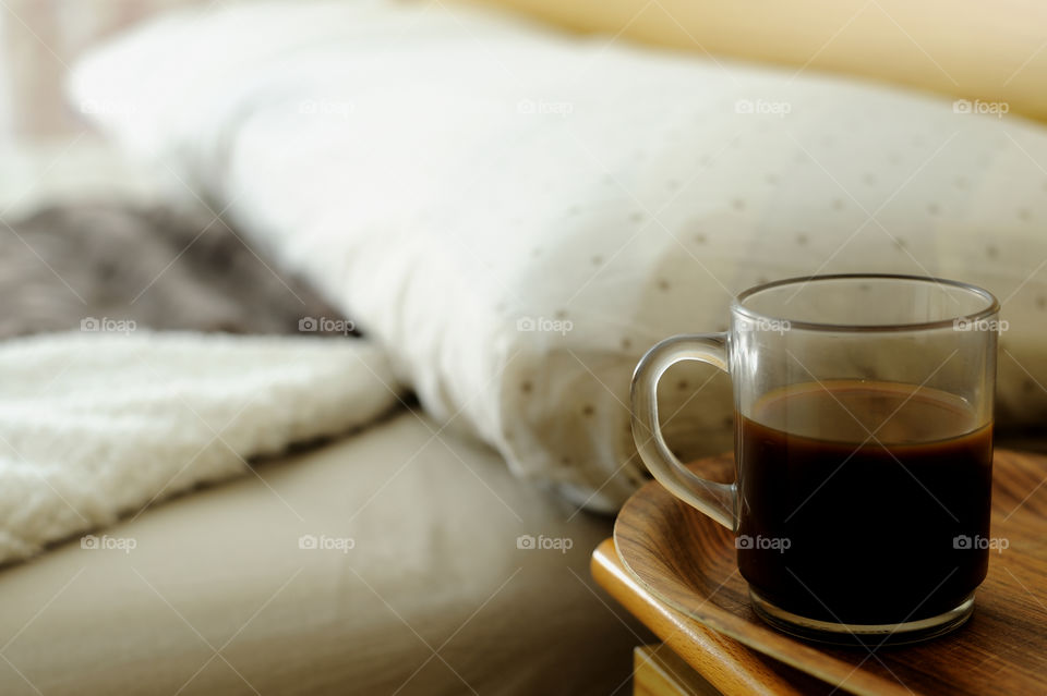Glass cup with coffee on a table next to an unmade bed.