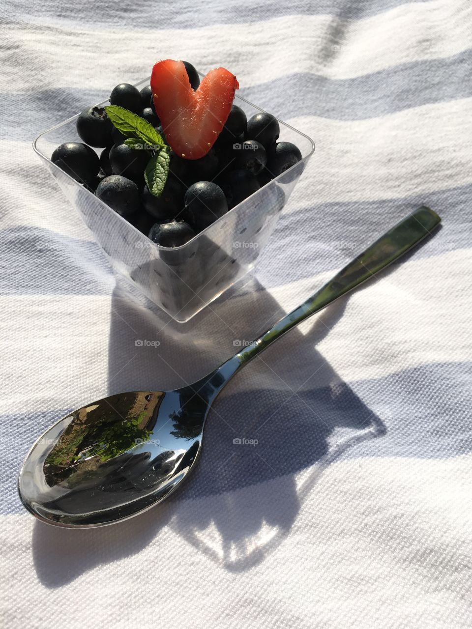 Square bowl full of blueberries with a heart shaped strawberry,sprig of mint and a spoon, outside in summer
