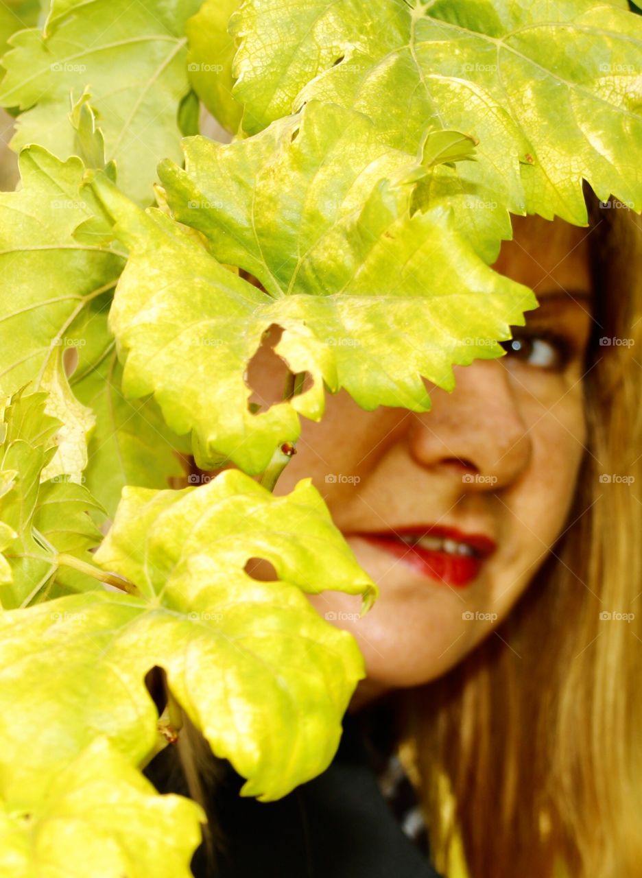 The model is showing her face behind some green leaves. The leaves of the plants are bright green and their texture is very smooth.