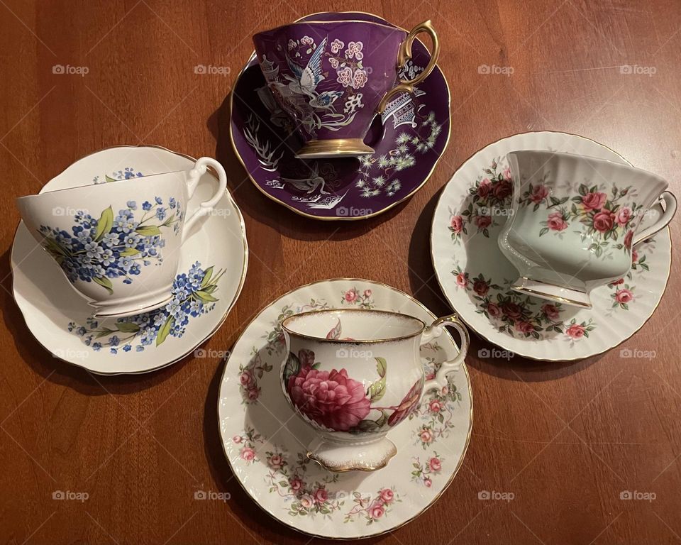Four antique teacups and saucers, each with a distinct, vintage charm. Each teacup features unique delicate patterns—including intricate floral motifs and gold detailing. The saucers are equally elegant, each matching the teacup in color and style.