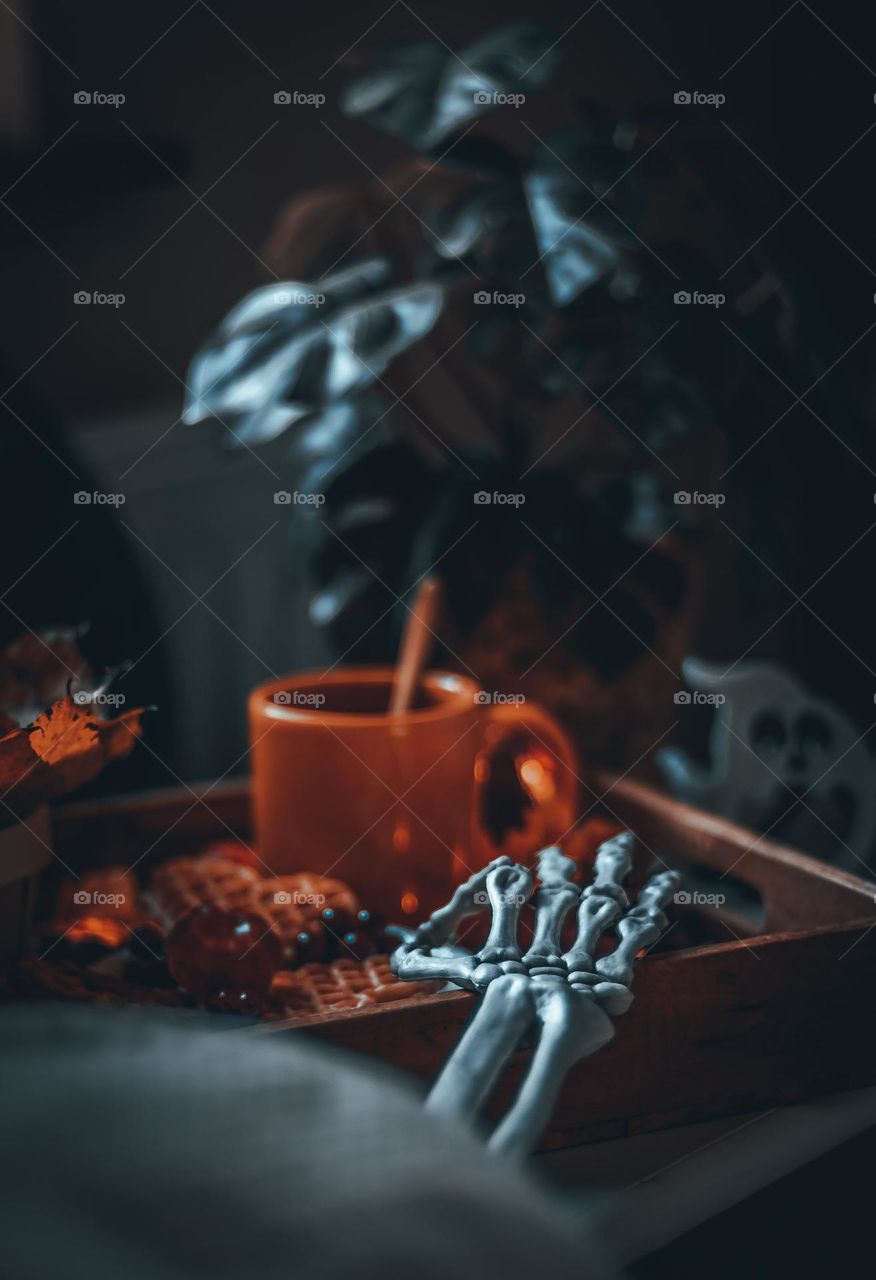 Skeleton hand reaching out from under the blanket on the bed at breakfast in a wooden tray standing on the bedside table with a mug of coffee, waffles, ghost glasses and autumn leaves garland, close-up side view. Halloween breakfast concept