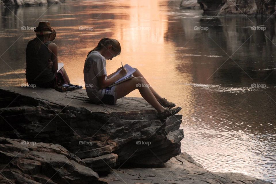 Teenage girls sitting on rock studying near lake