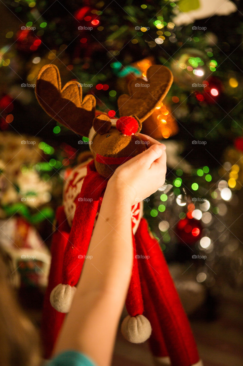 My girl loves this little Christmas reindeer. Image of girl's hand holding face of Christmas reindeer with Christmas tree and lights in background.