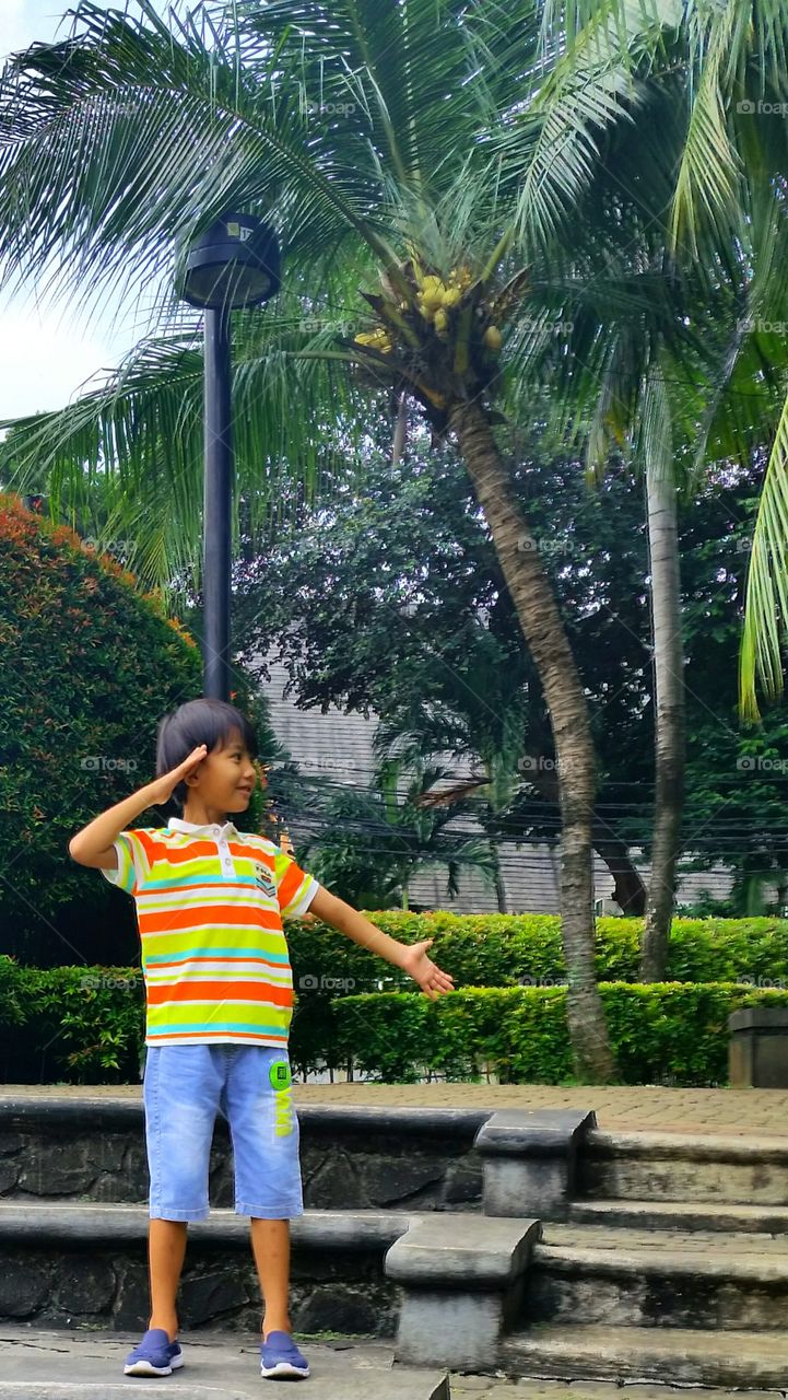 A boy standing in the park. The park has a lot plants and tree like coconut tree.