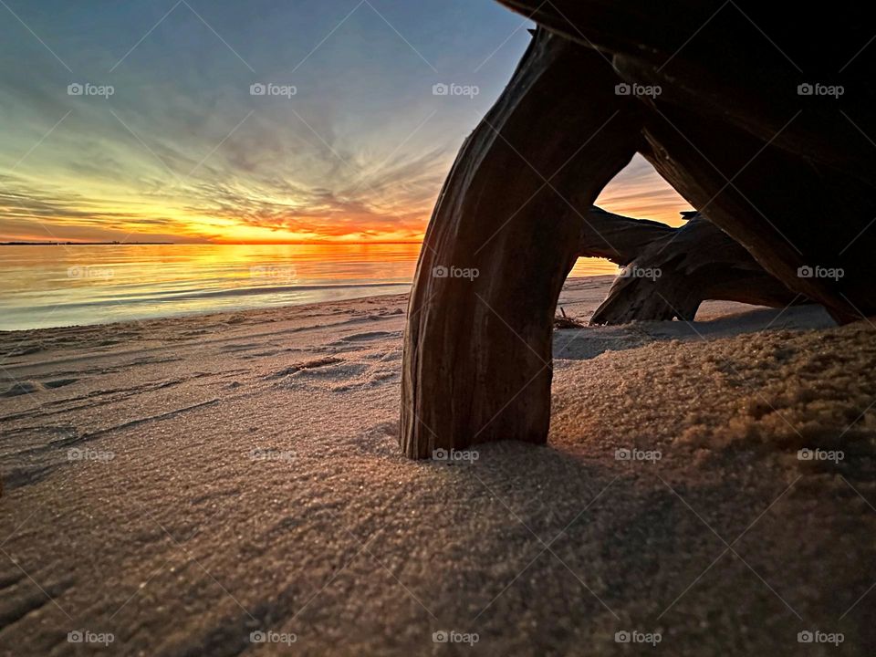 A descending sunset reflection off the waters surface. Taken from underneath and around a buried log in the sugary white crystal sand on the beach