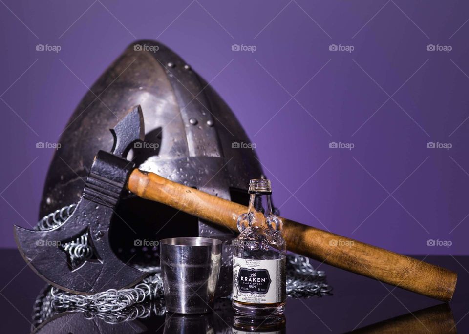 medieval themed image with miniature bottle of liquor and a metal chalice shot glass placed in front of a metal helmet, metal chainmail, and an axe