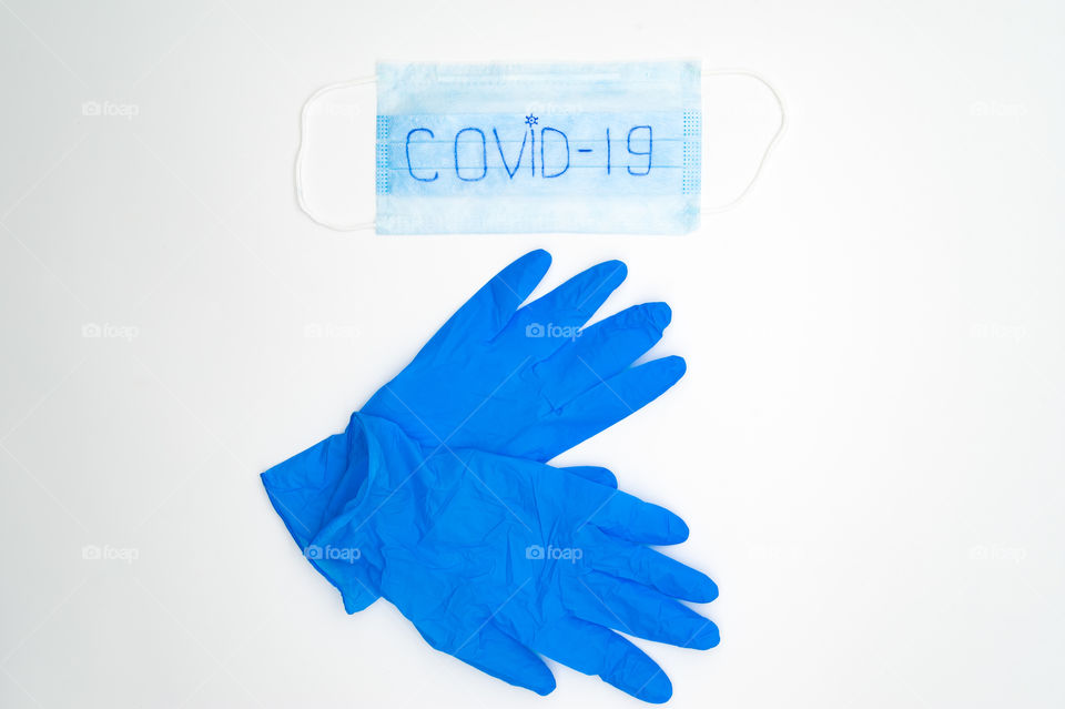 On a white background lies a blue medical mask and blue rubber gloves.  The inscription COVID-19 on the mask