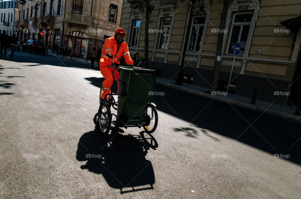Clean worker in a bright orange uniform rides on a special bicycle with garbage box along the street. Tbilisi, Georgia.