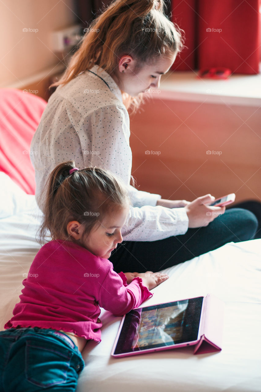 Teenage girl using mobile phone together with her little sister watching animated movie on tablet, both girls sitting in bed in bedroom