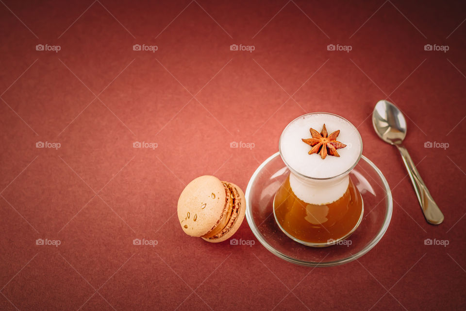 Some beverage with coffee, milk and spice in a glass near the chocolate macaron on the brown background.