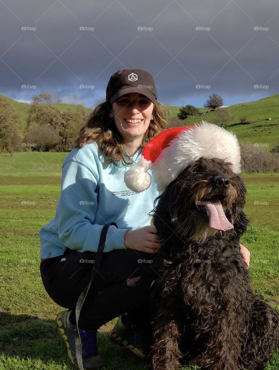 A girl and her dog pose for a holiday photo with a Santa hat while on a walk in nature. 