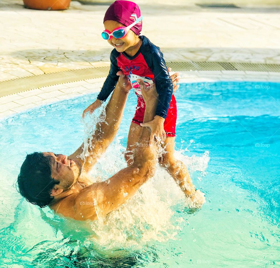 A father and daughter enjoying a lot at swimming pool in summertime,father holds his daughter upright position and daughter laughs loudly.