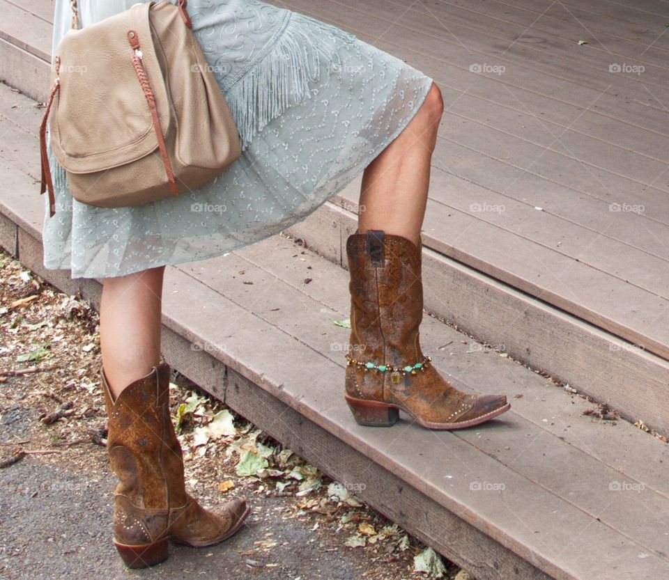 A cowgirl stands with one boot on a stair while she reviews hanging art. 