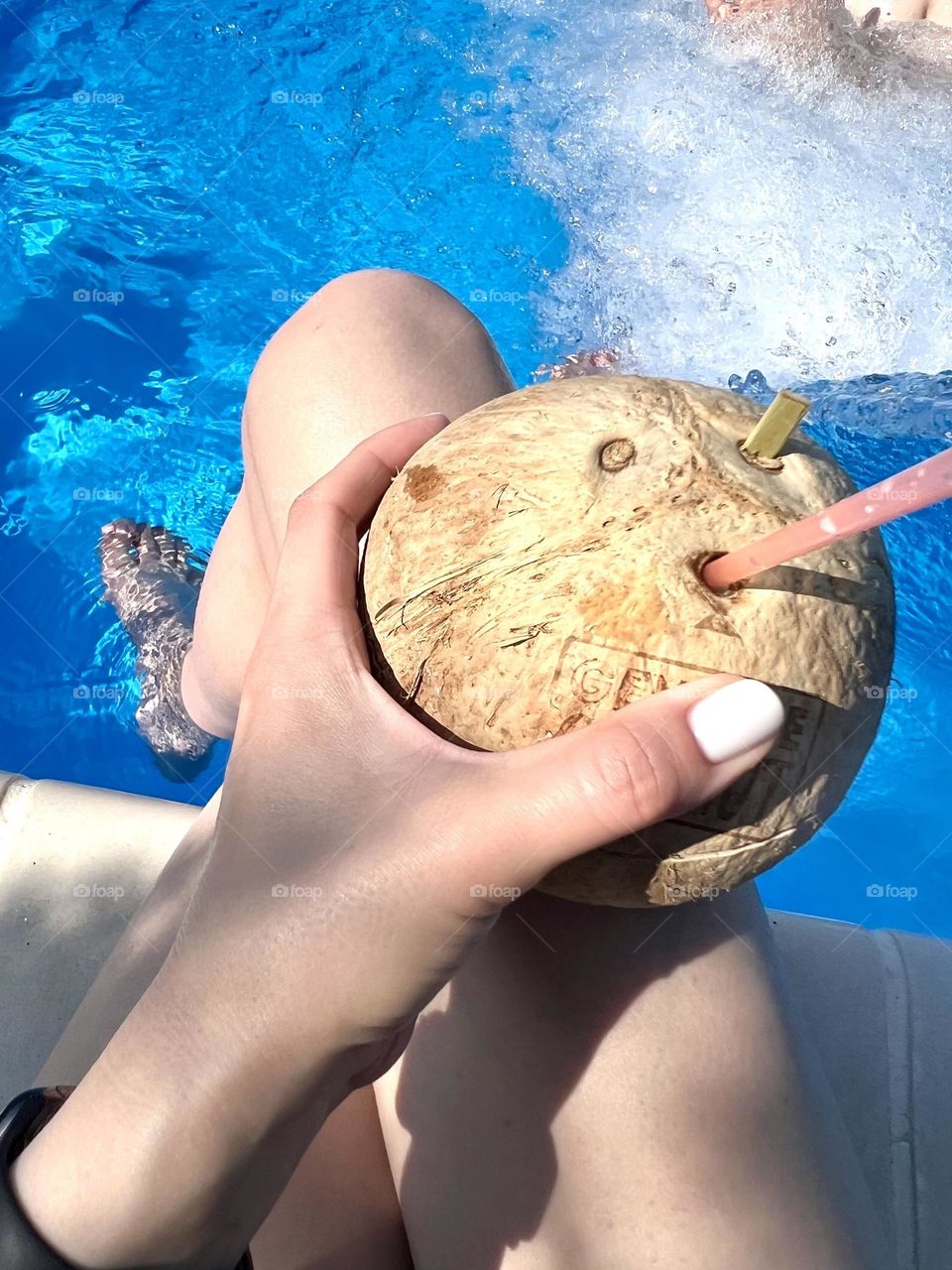 Сверху неузнаваемая девушка сидит с кокосовым коктейлем  возле синего бассейна 