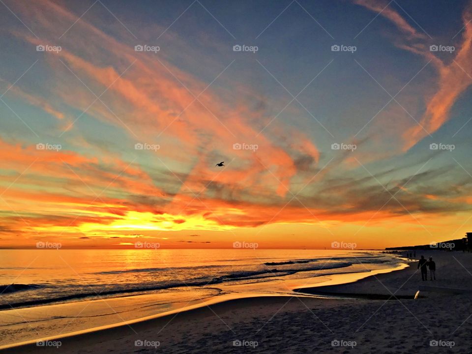 Silhouette of a couple walking along the sandy beach toward the last light of the day, as it descends below the horizon. 