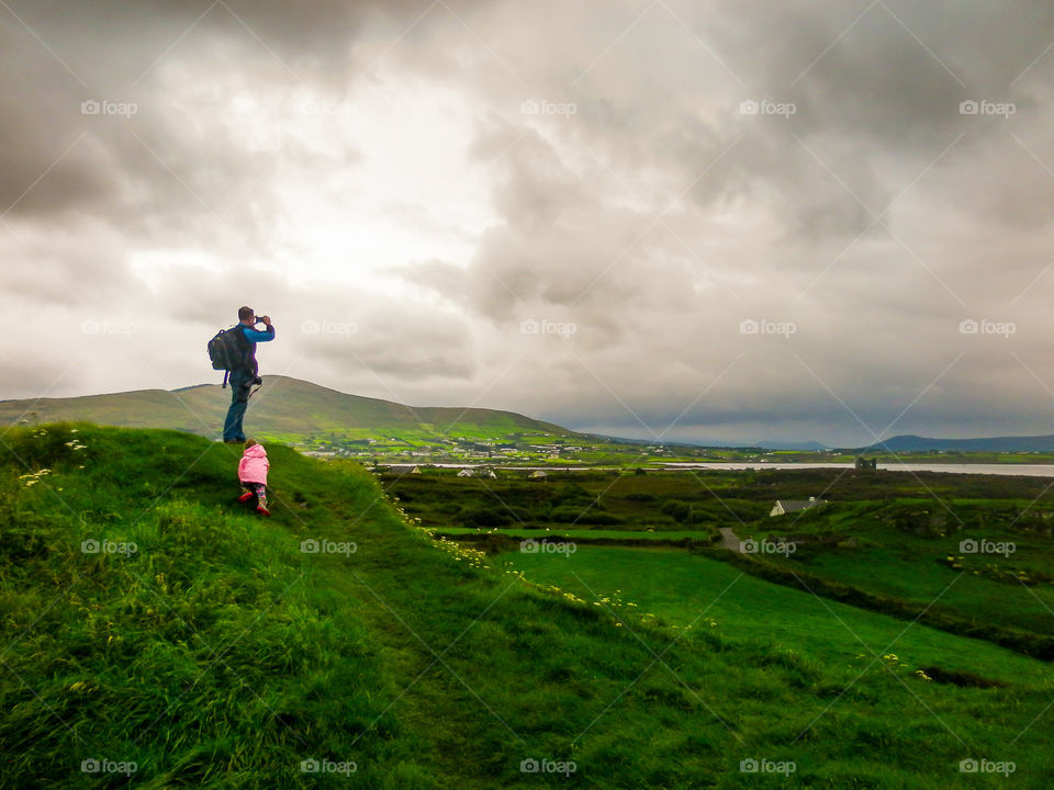 Such a beautiful memory - dad and daughter sightseeing on a cloudy cold day in Europe. Image of dad taking photo of green scenery with grey clouds and little girl in pink jacket