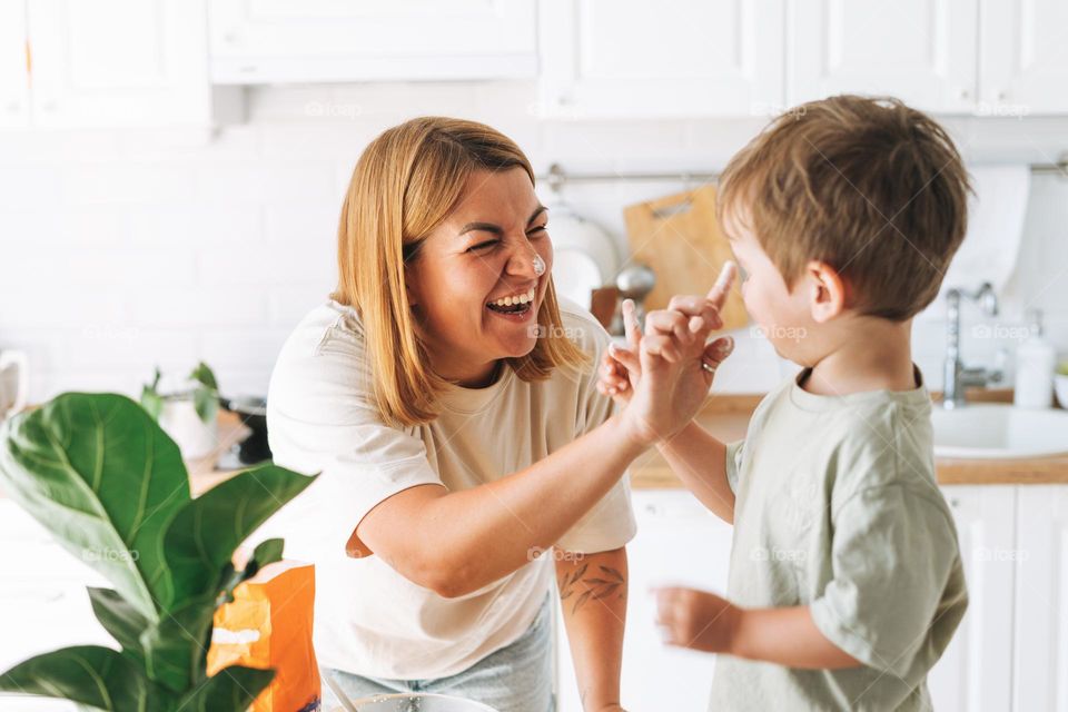 Young woman mother and her toddler boy son have fun while cooking with flour at table in bright kitchen at home