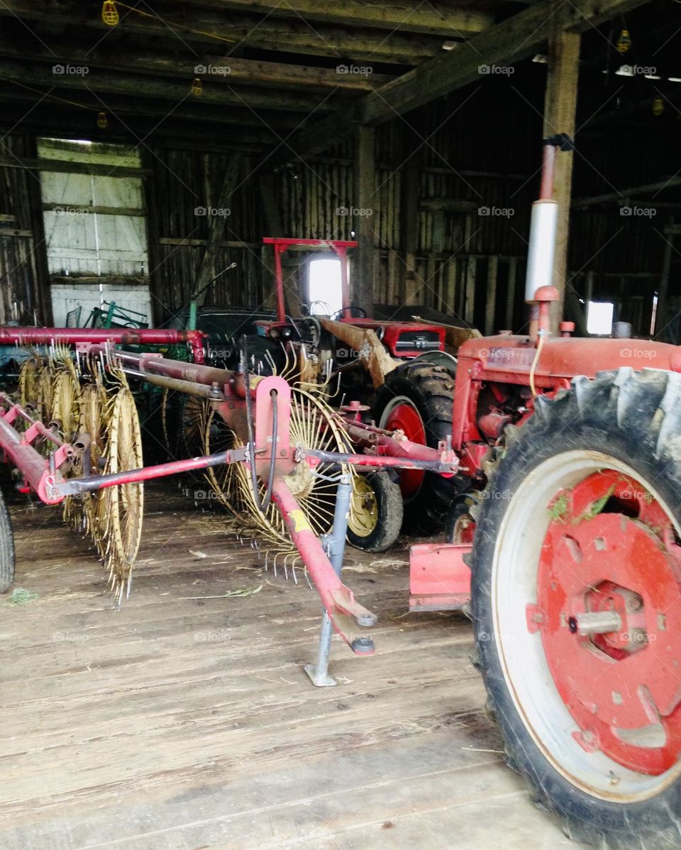 In an open barn, old red farming equipment sit quietly, awaiting the call to the fields. The rusted but sturdy machinery, a mix of tractors, plows, and harvesters, are lined up in neat rows. Their weathered paint still hold a nostalgic charm.