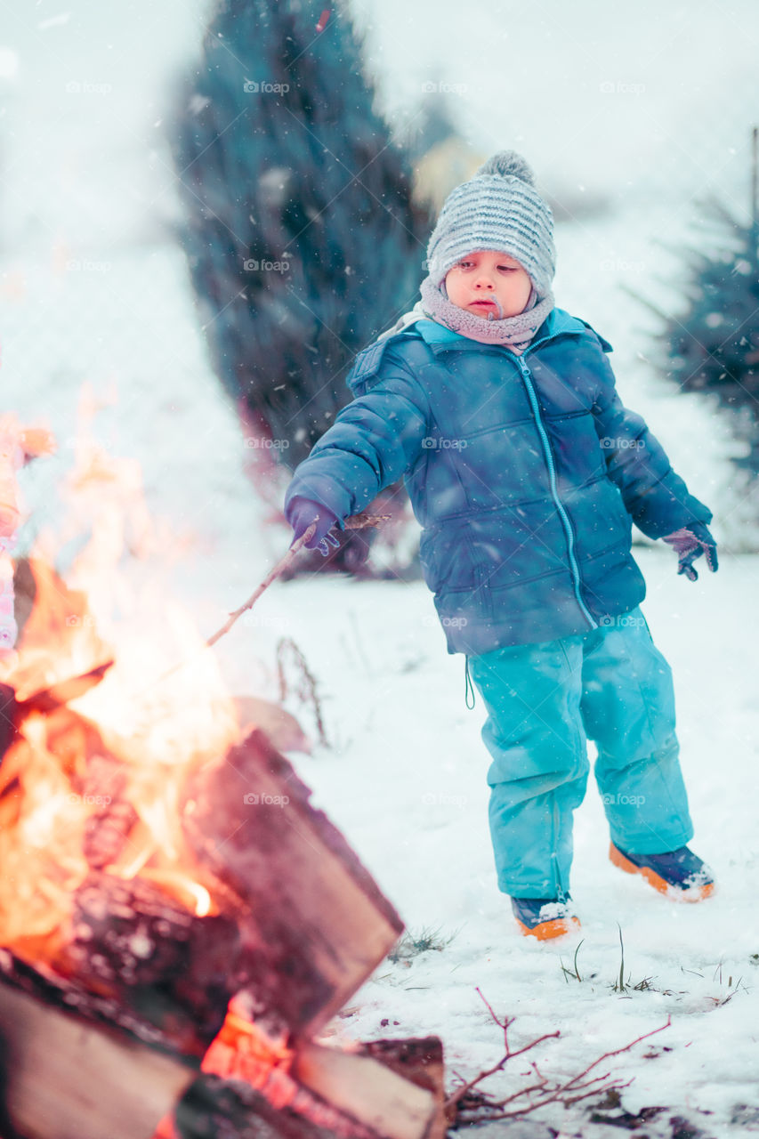 Boy playing a stick by campfire outdoors in the winter. Kid wearing warm clothes, scarf and wool cap