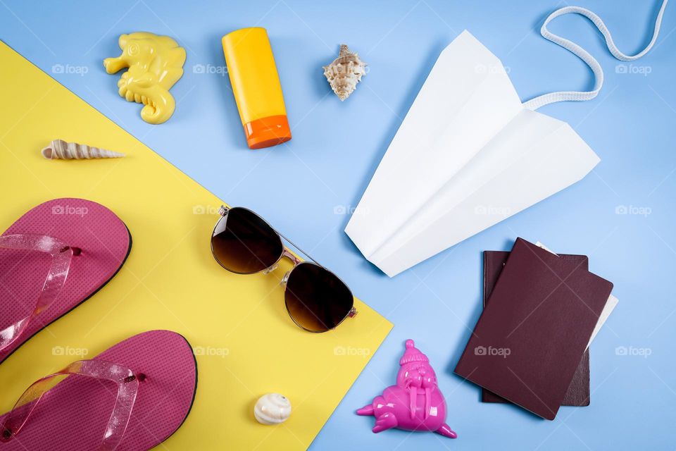 Sunglasses, sunblock, flip-flops, passports, paper airplane and seashells lie on a blue-yellow background, flat lay close-up.