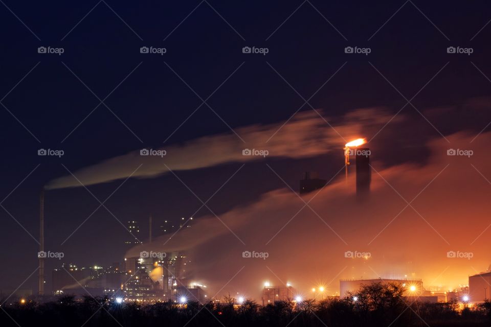 An oil refinery at night with smoke billowing from tall chimneys.