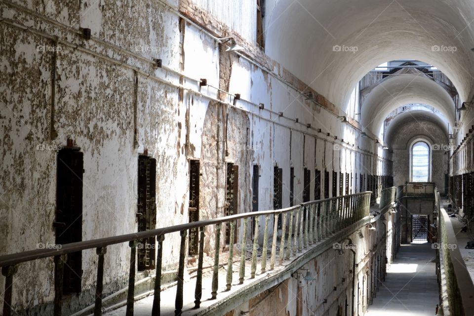 A second floor hallway in a neglected, abandoned prison with a metal guardrail and a series of doors