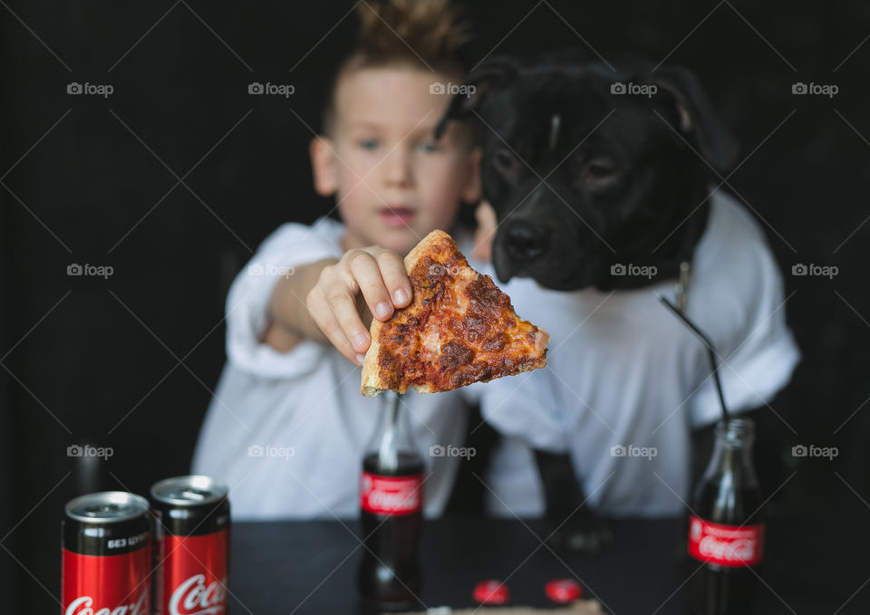 cute kid with his friend dog, dressed in white T-shirts, sitting at the table, eating pizza and drinking Coca-Cola. Emotions, joy. Black background