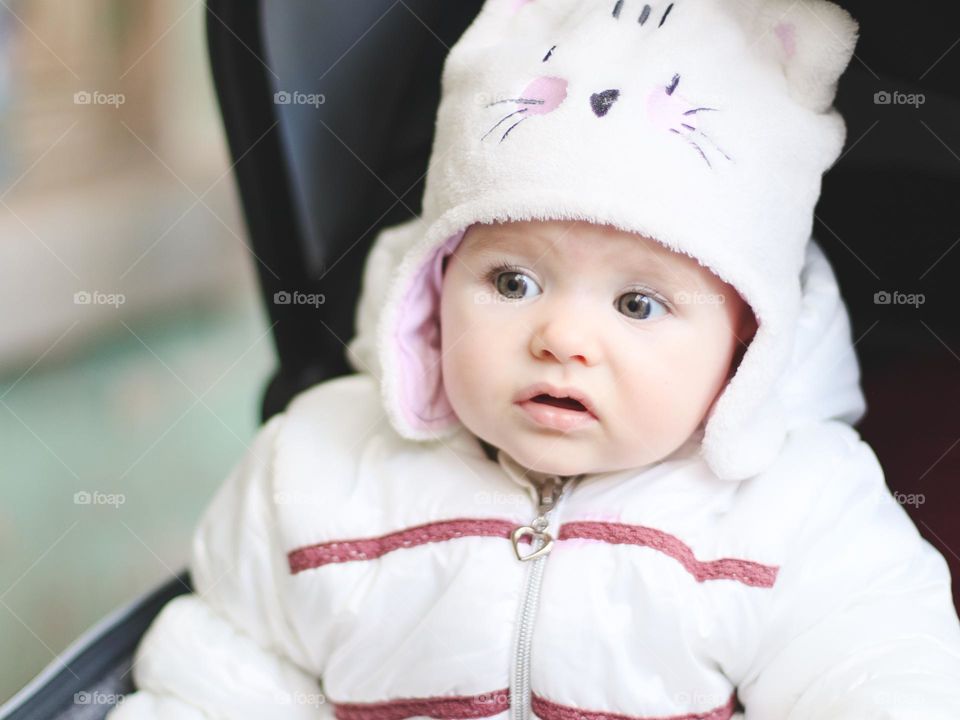 Portrait of a beautiful little Caucasian baby girl in a winter hat, jacket with a very surprised and curious emotion on her face sits in a stroller and looks to the side, close-up side view. Surprised emotion concept,