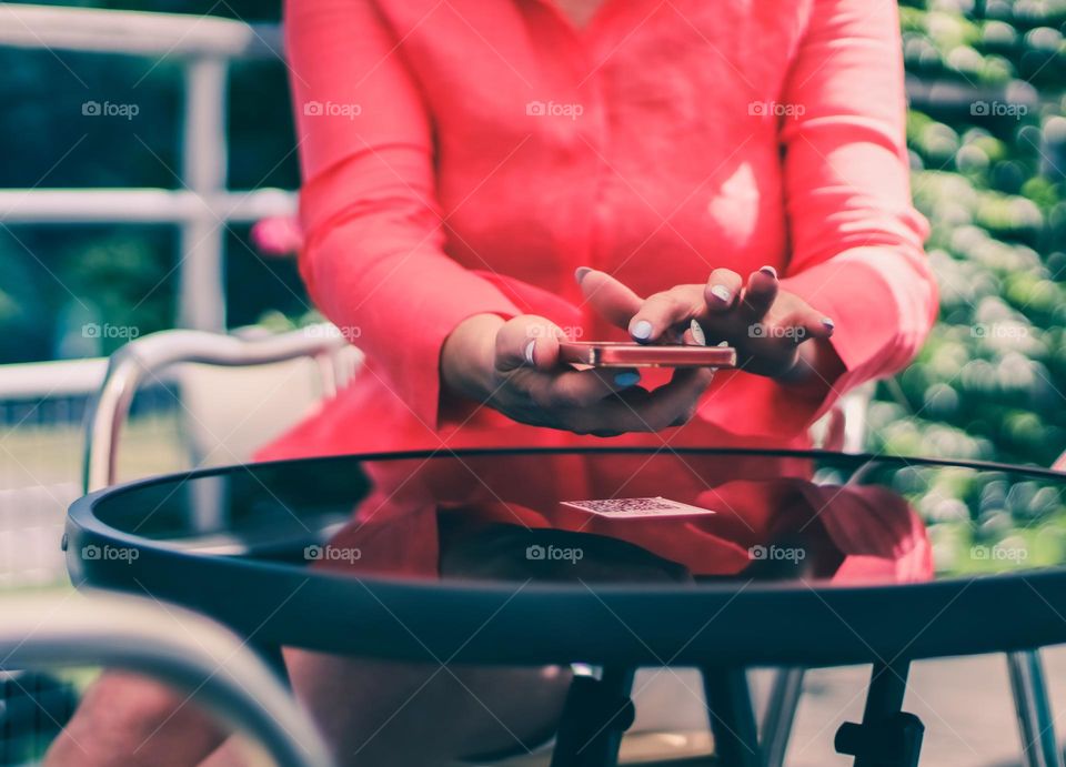 The hands of a young caucasian woman in a red shirt hold a mobile phone in her hands and scan the barcode of the menu on a round black glass table of a street cafe, vtd side close-up. The concept of using a smartphone.