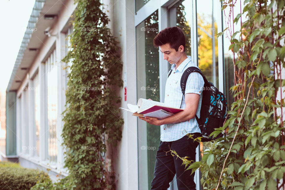 Student holding a notebook and carrying a backpack standing at the front of university building. Young boy wearing blue shirt and dark jeans