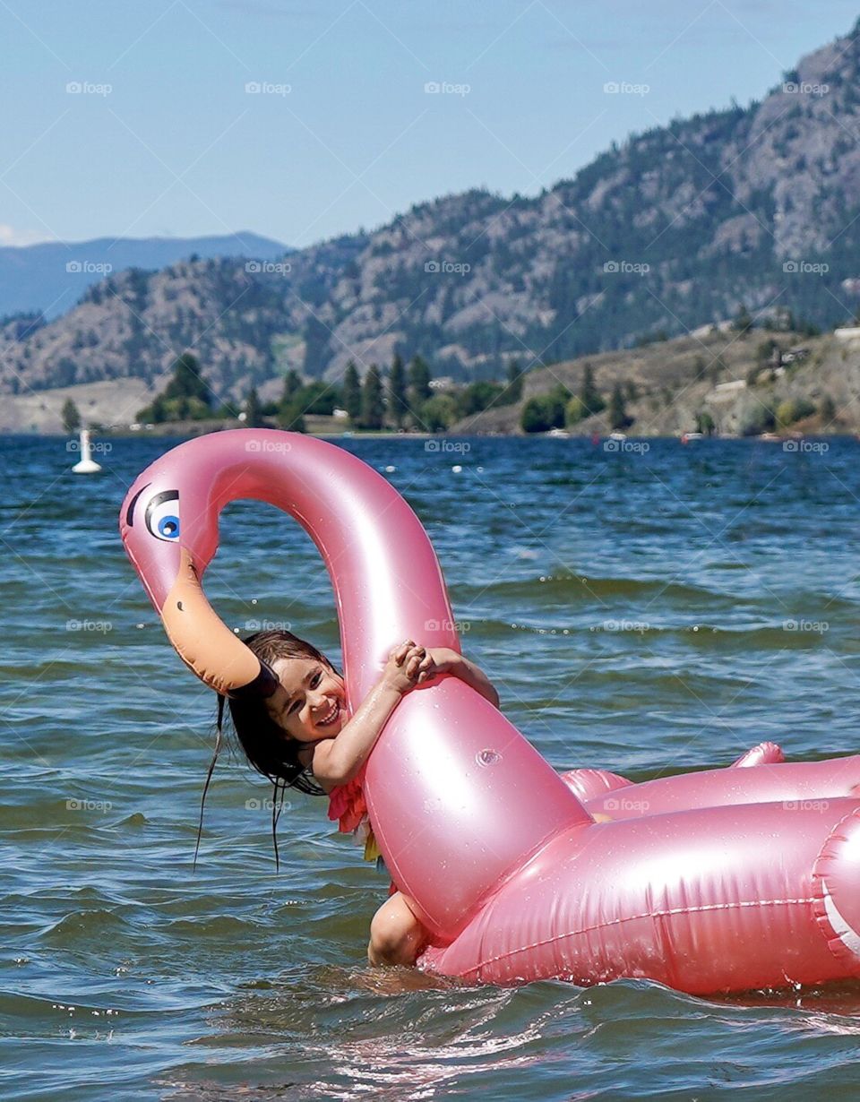 Child hanging onto a pink flamingo blowup toy in the middle of a sunny lake