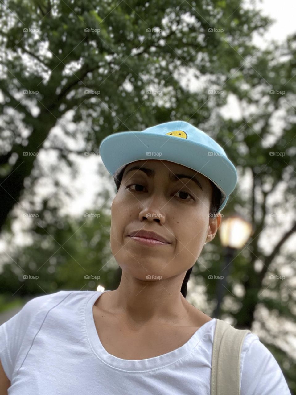 Young adult woman selfie portrait wearing blue baseball hat with trees backdrop. 